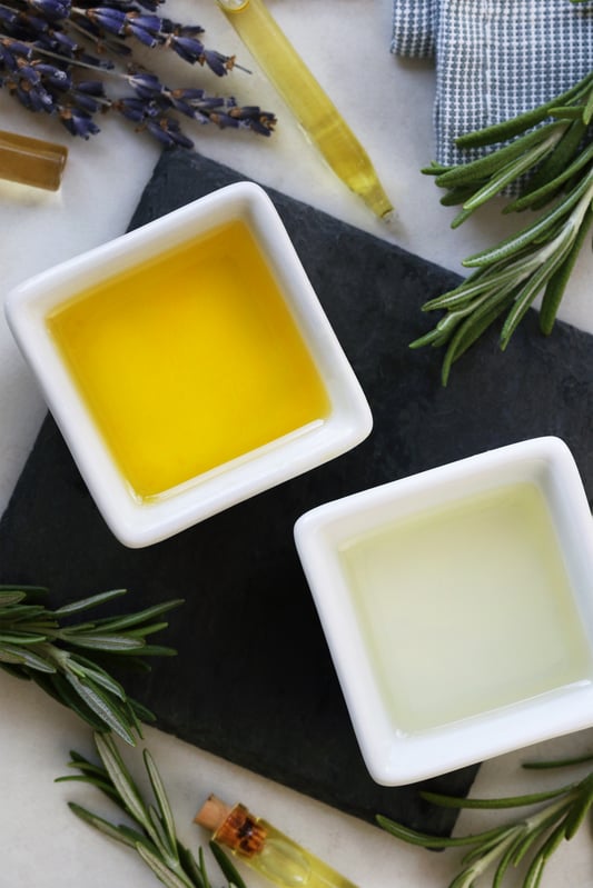 Vibrant deep yellow jojoba oil and clear argan oil arranged with fresh rosemary and lavender flowers to be combined into a DIY hair moisturizer.