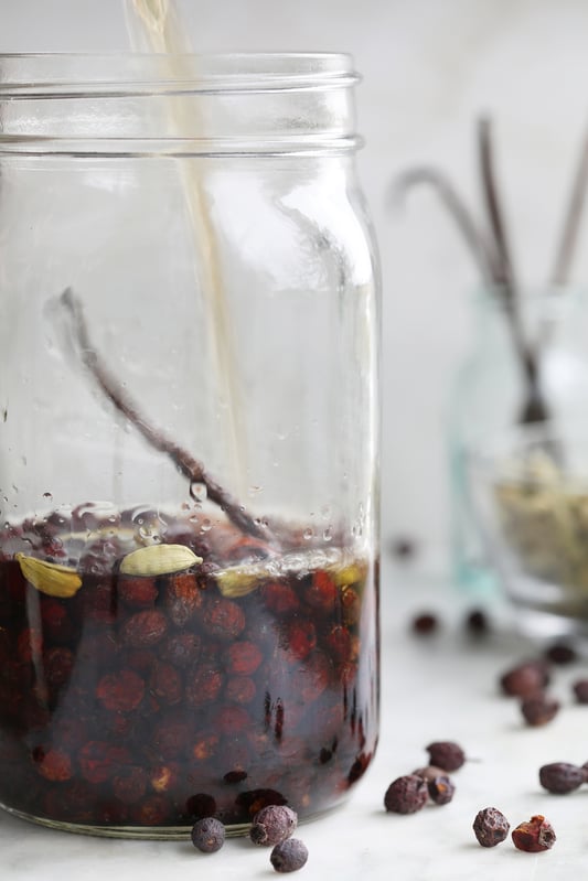 Mason jar filled with dried dark read hawthorn berries and cardamom pods with golden liquid being poured into it.