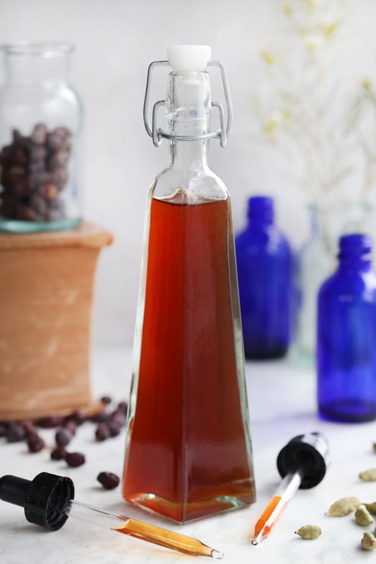 Unique shaped clear class bottle with rich red liquid surrounded by medicine droppers, dried berries and cardamom pods. 