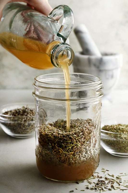 Pouring apple cider vinegar over lavender and rosemary herbs to create an infused herbal vinegar for cooking, cleaning, and skincare. A mason jar is full of herbs to be covered with apple cider vinegar for infusing. 