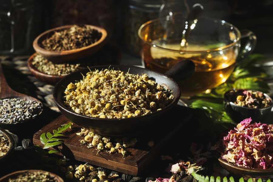 A large bowl of chamomile flowers sits in the center of many bowls of herbs and a glass of golden tea in the background.