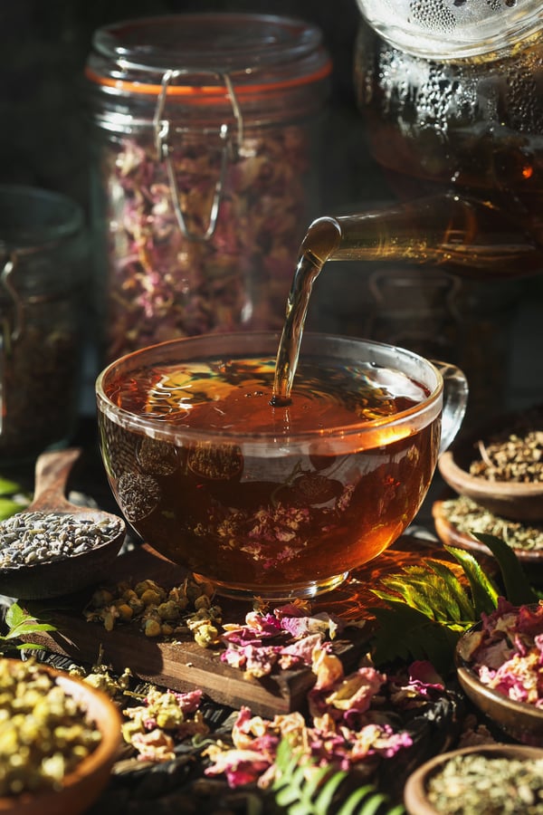 A hot golden colored tea is poured from a glass tea pot into a small glass teacup. The teacup is surrounded by many herbs included in the tea blend.