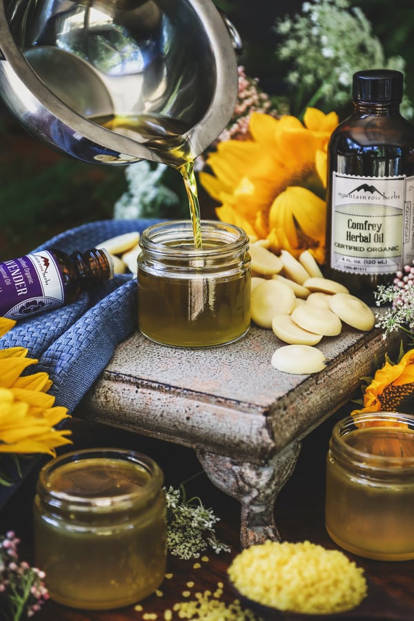 Melted beeswax salve is poured from a silver double boiler bowl into a small glass jar. Jar is surrounded by Comfrey Herbal Oil and Lavender Essential Oil bottles.