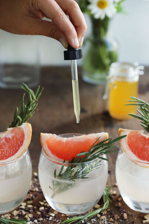 Bitter sodas garnished with slices of fresh grapefruit and sprigs of rosemary. A dropper of homemade bitter releasing drops into the bubbly water is held over a glass.
