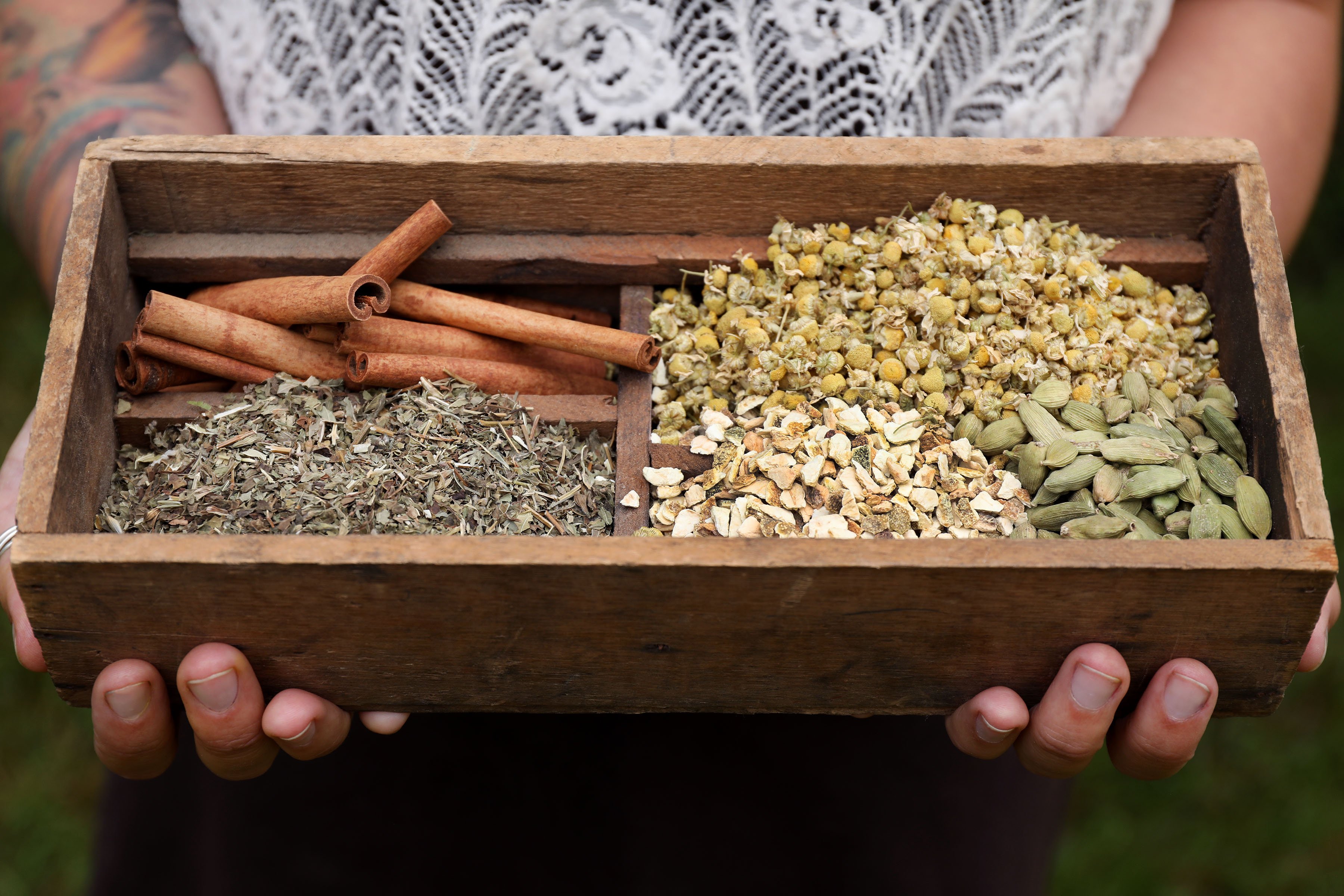 A wooden box filled with dried herbs including cinnamon sticks, chamomile flowers, cardamom pods, and dried roots and leaves. 