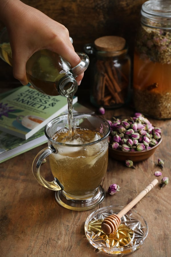 Herbal marshmallow tea being poured into a clear mug with ice cubes. A plate of honey and a bowl of rose buds are featured, along with jars of tincturing herbs and books on herbalism. 