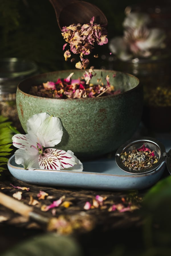 Pink rose petals are sprinkled into a bowl containing the tea blend mentioned in this blog. The bowl is green and is sitting on a blue tray with flowers and herbs.