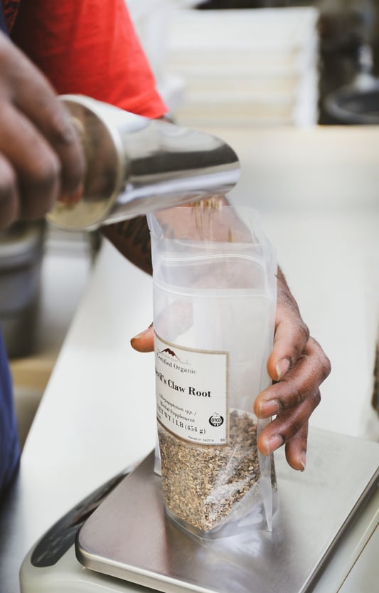 Hand using metal scoop to pour herbs into bag on scale