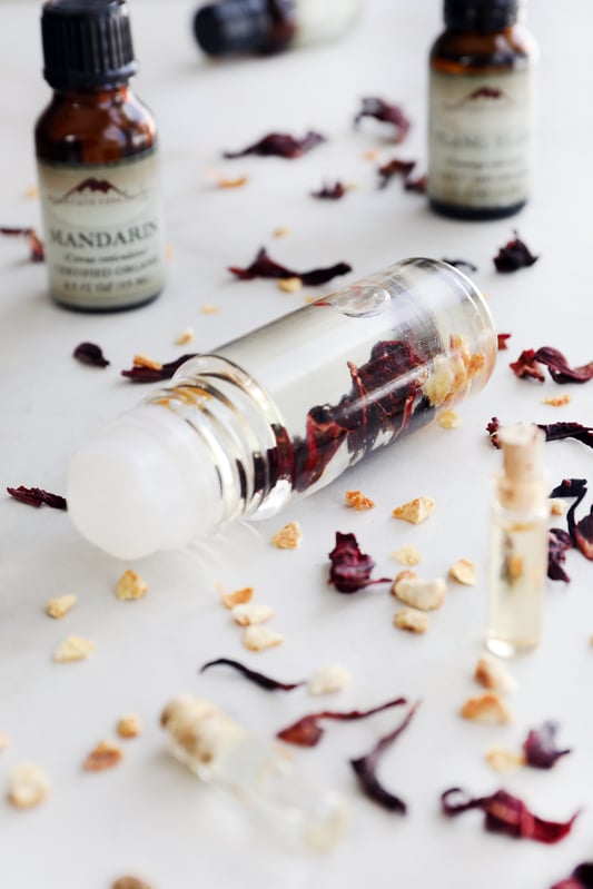 Clear glass roll top perfume bottle filled with rich red hibiscus flowers surrounded by essential oil bottles and botanicals.