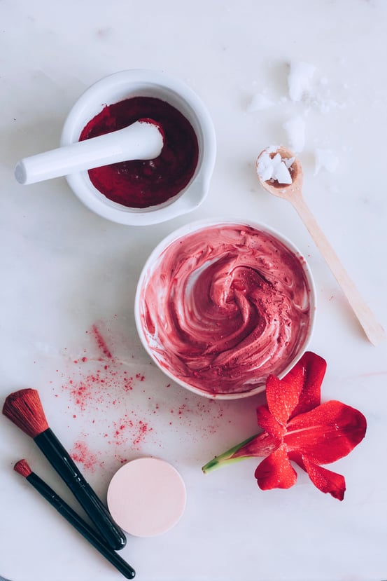 Bowl of red tinted cream surrounded by mortar and pestle with makeup brushes and homemade makeup and hibiscus flower