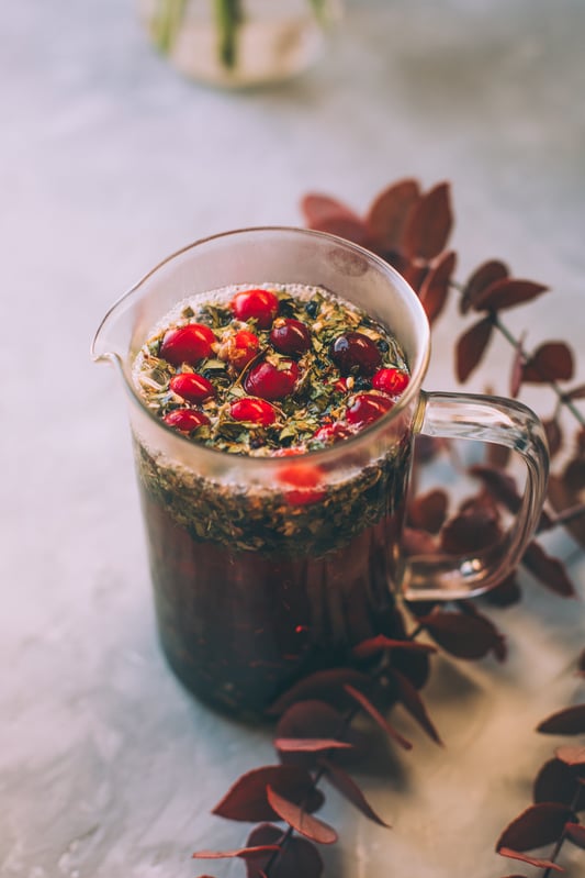 A pitcher of holiday cocktail, or mocktail without alcohol, made with organic maqui berries and bilberries in trendy glass jars with gold rims and garnished with pomegranate seeds, fresh lime wheels, and cranberries.