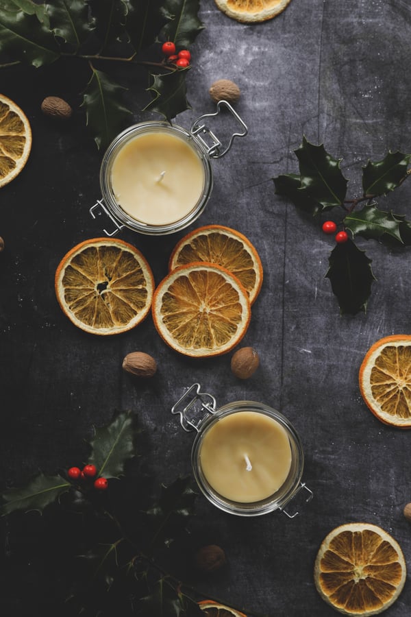 Homemade spiced cream beeswax candles in pantry jars on a table surrounded by dried orange slices and holly with berries