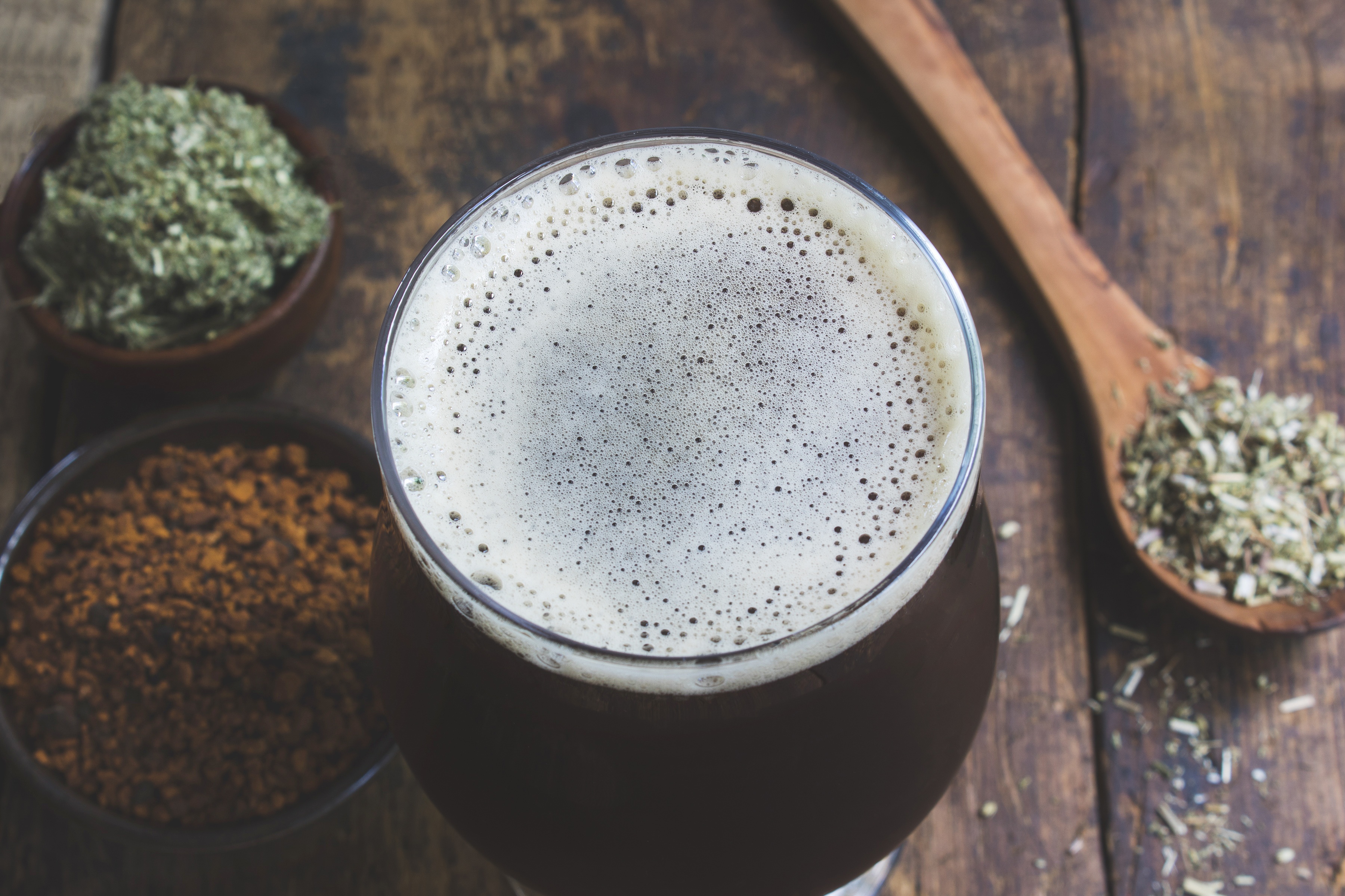 Glass with deep roots gruit herbal beer filled to top with foam and herbs on the table near the sides