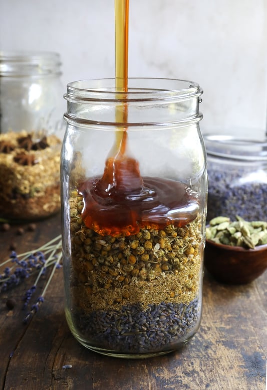 Pouring honey into mason jar filled with colorful dried herbs in it