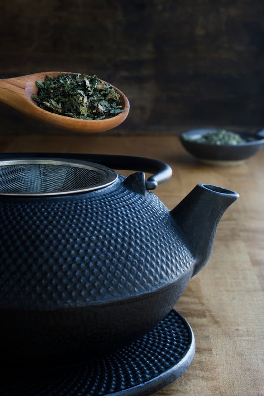 Black cast iron tea pot being illed with green herbs on wooden table