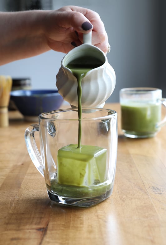 Hand using pitcher to pour matcha tea over ice cube in glass tea cup