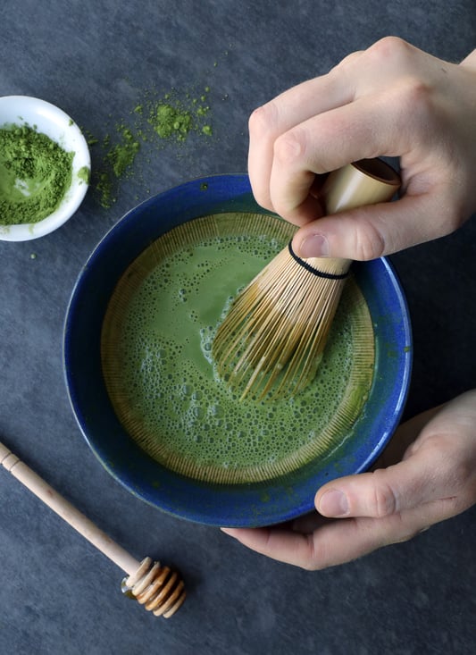Hands using matcha whisk mixing matcha powder with liquid in a blue ceramic bowl