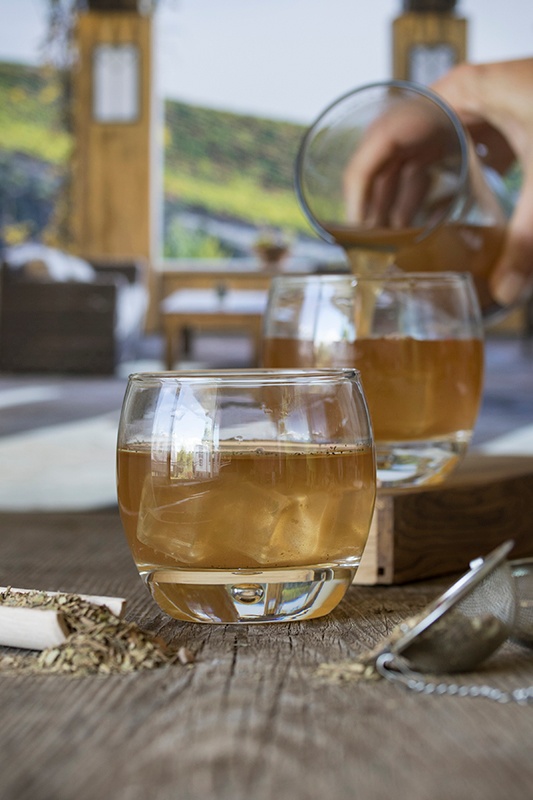 Pouring iced mint chocolate mate tea into round class on wooden counter with loose leaf tea and strainer