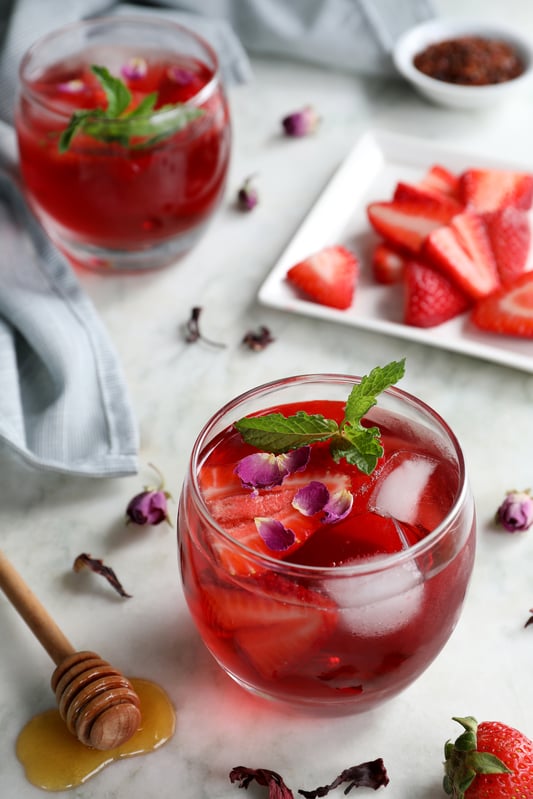 Small glass tumblers are filled with bright red herbal iced tea and garnished with fresh strawberry slices, mint leaves, and rose petals. Arranged on a white marble counter top with a plate of fresh strawberries and honey dripping from a scoop.