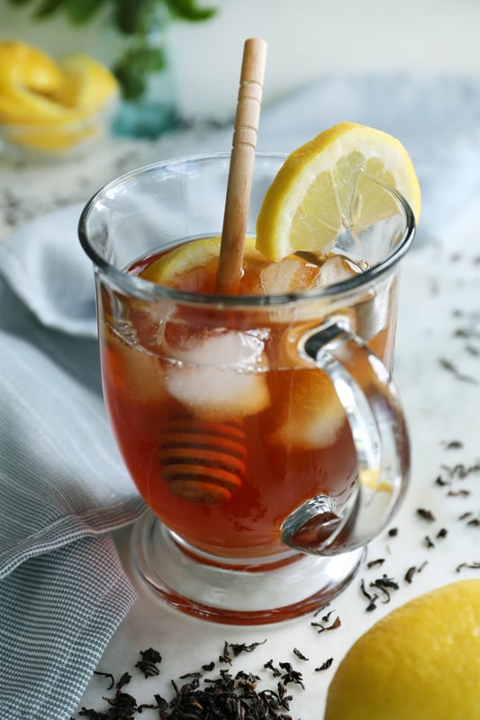 A clear glass mug is full of iced black tea, fresh lemon wedges, and a honey scoop. Arranged on a white marble counter with fresh lemons and loose leaf black tea.