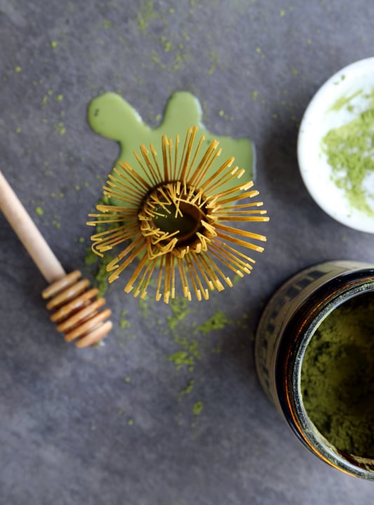 Matcha whisk facing upward towards ceiling near matcha powder and honey 