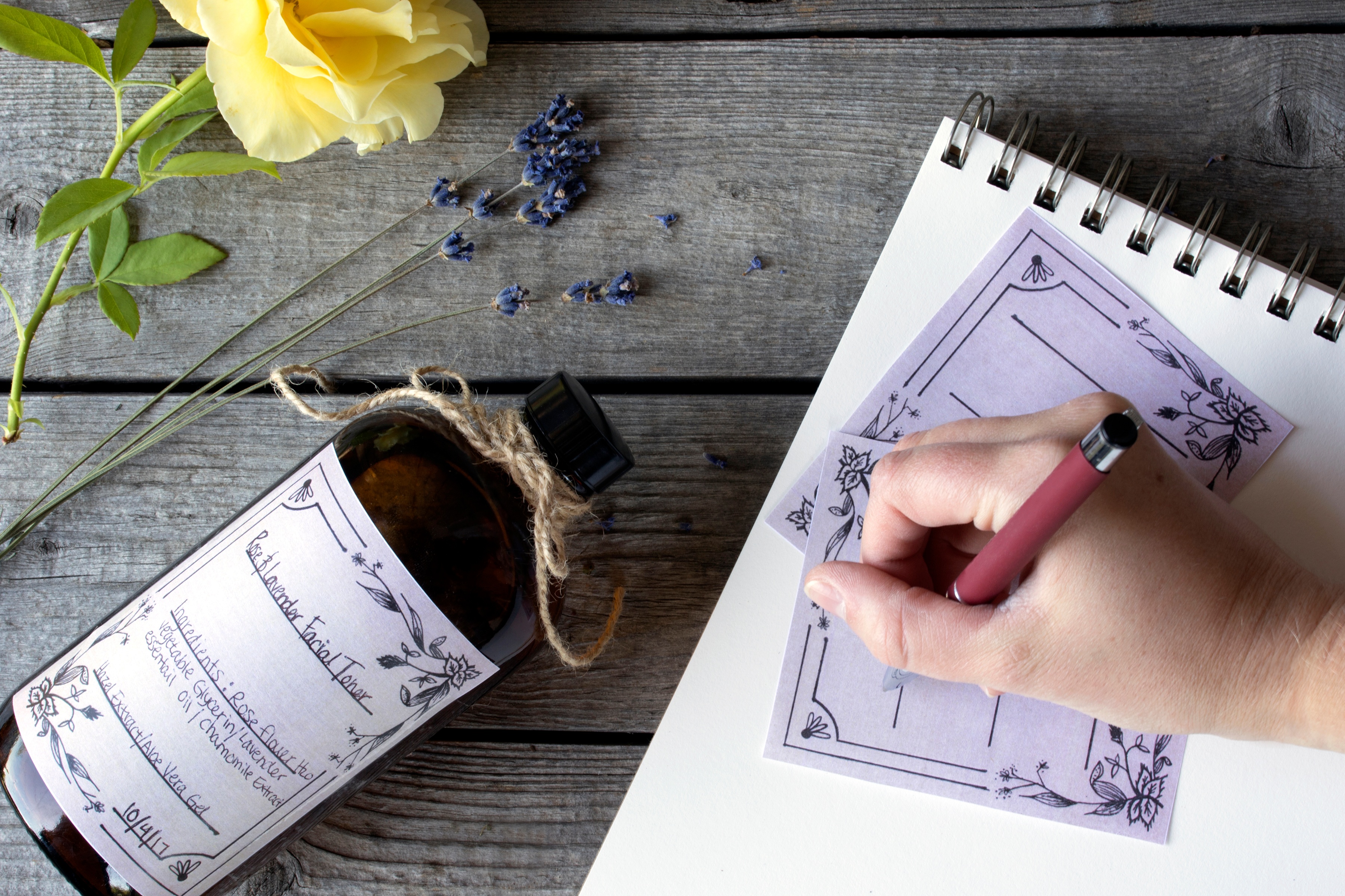 Person handwriting on a printed label with a yellow rose and lavender flowers on a wooden table.