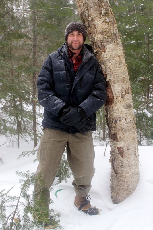 Handsome young man in snow gear leaning against tree in a snowy forest while pursuing organic, wildharvested chaga mushrooms.