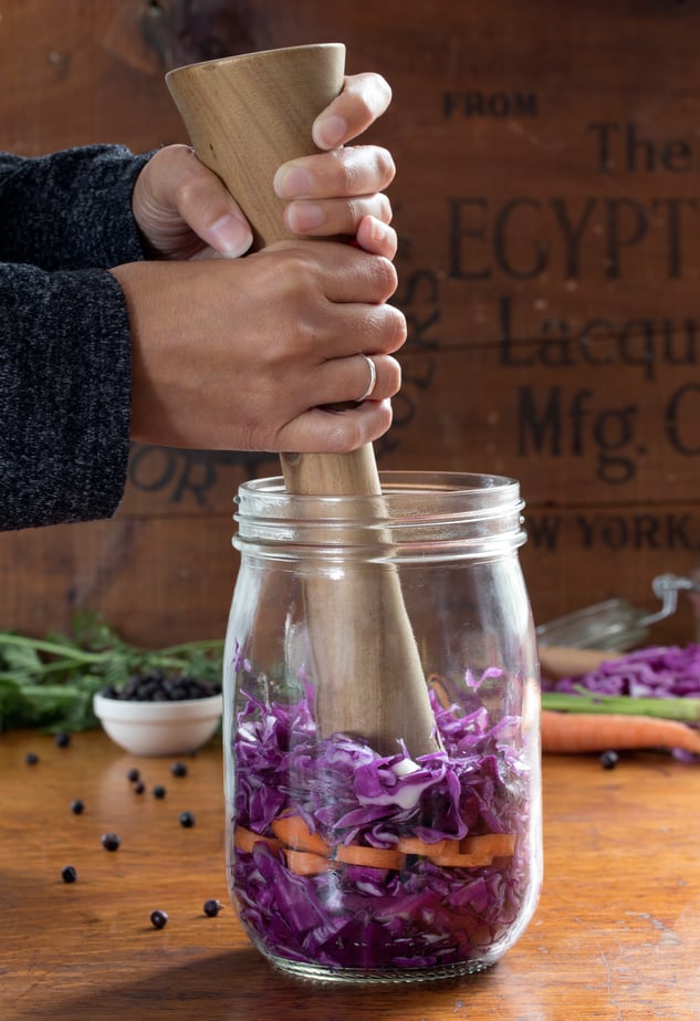 Hands using wooden pestle to massage cabbage to make sauerkraut in a glass jar