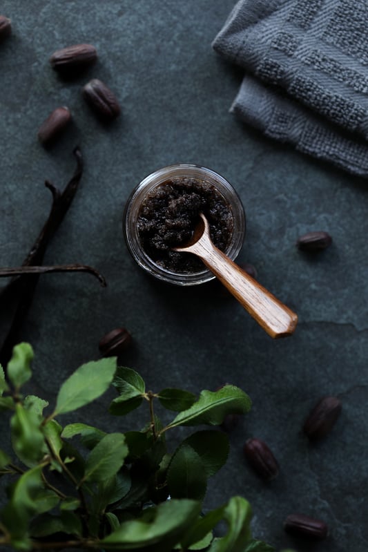 A DIY body scrub with vanilla and jojoba meal, honey, and coarse sea salt in a small glass salve jar. A wooden spoon is sitting in the salt scrub. Fresh jojoba beans are scattered around. 