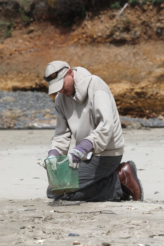 Jonathan kneeling on sand with bucket to pick up trash on the beach while helping with volunteer work