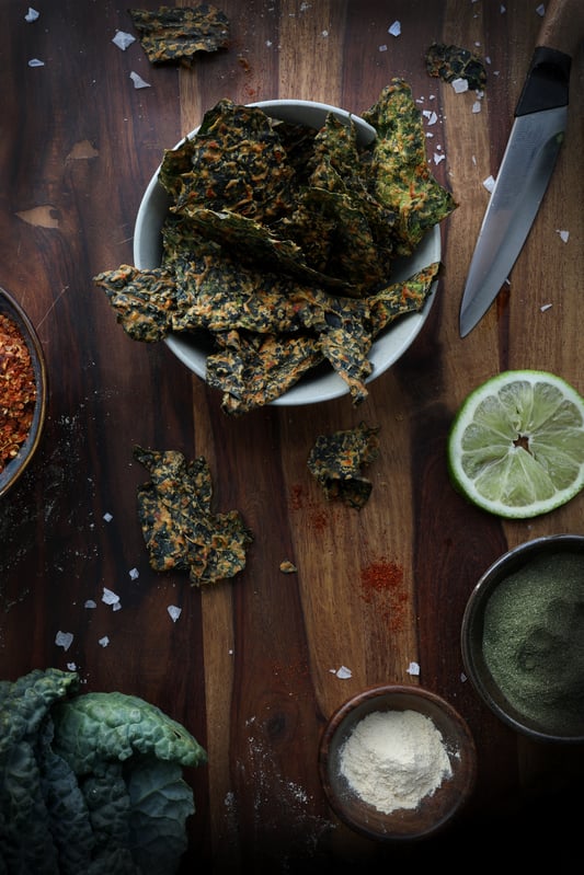 A ceramic bowl filled to the brim with spicy kale chips. Surrounding the bowl are various ingredients, fresh lime, chili flakes, fresh kale, and powders, all on a wooden cutting board.