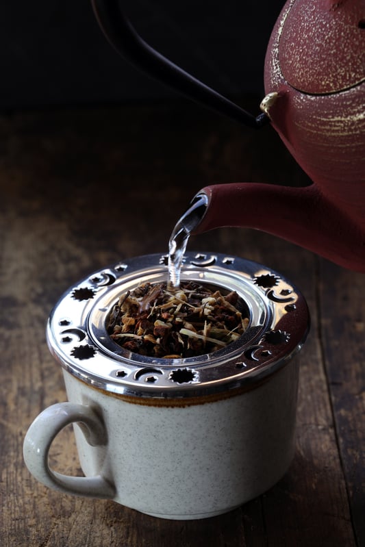 Tea kettle pouring hot water into mug holding celestial tea strainer filled with loose leaf tea