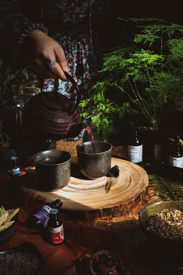Someone in a blue patterned dress pouring a red tea into a steaming cup surrounded by plants and herbal products.