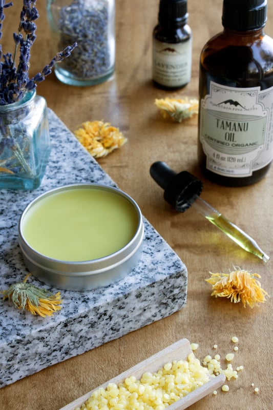 Homemade skin salve in tin surrounded by beeswax pastilles in wooden scoop, dried calendula flowers, lavender, and tamanu oil bottle on marble slab