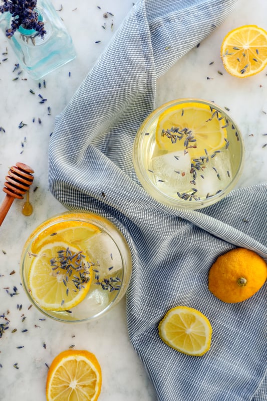 Lemon lavender sodas garnished with fresh slices of citrus fruit and lavender flowers are arranged with honey and slices of lemons and oranges on a white marble counter top.