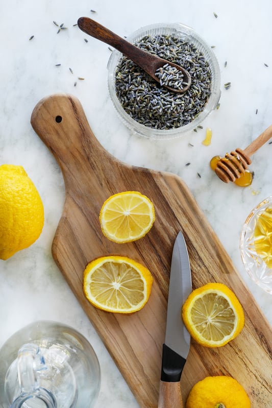 Ingredients for a lemon lavender soda are arranged with a wooden cutting board on a white marble countertop. Honey, fresh lemons, lavender flowers alongside a knife and honey scooper.