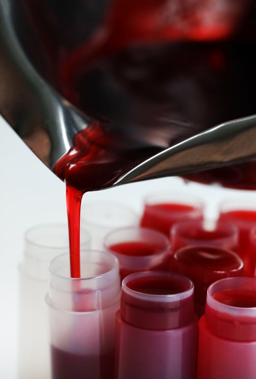 Steel pitcher pouring red lip balm liquid into plastic lip balm tubes