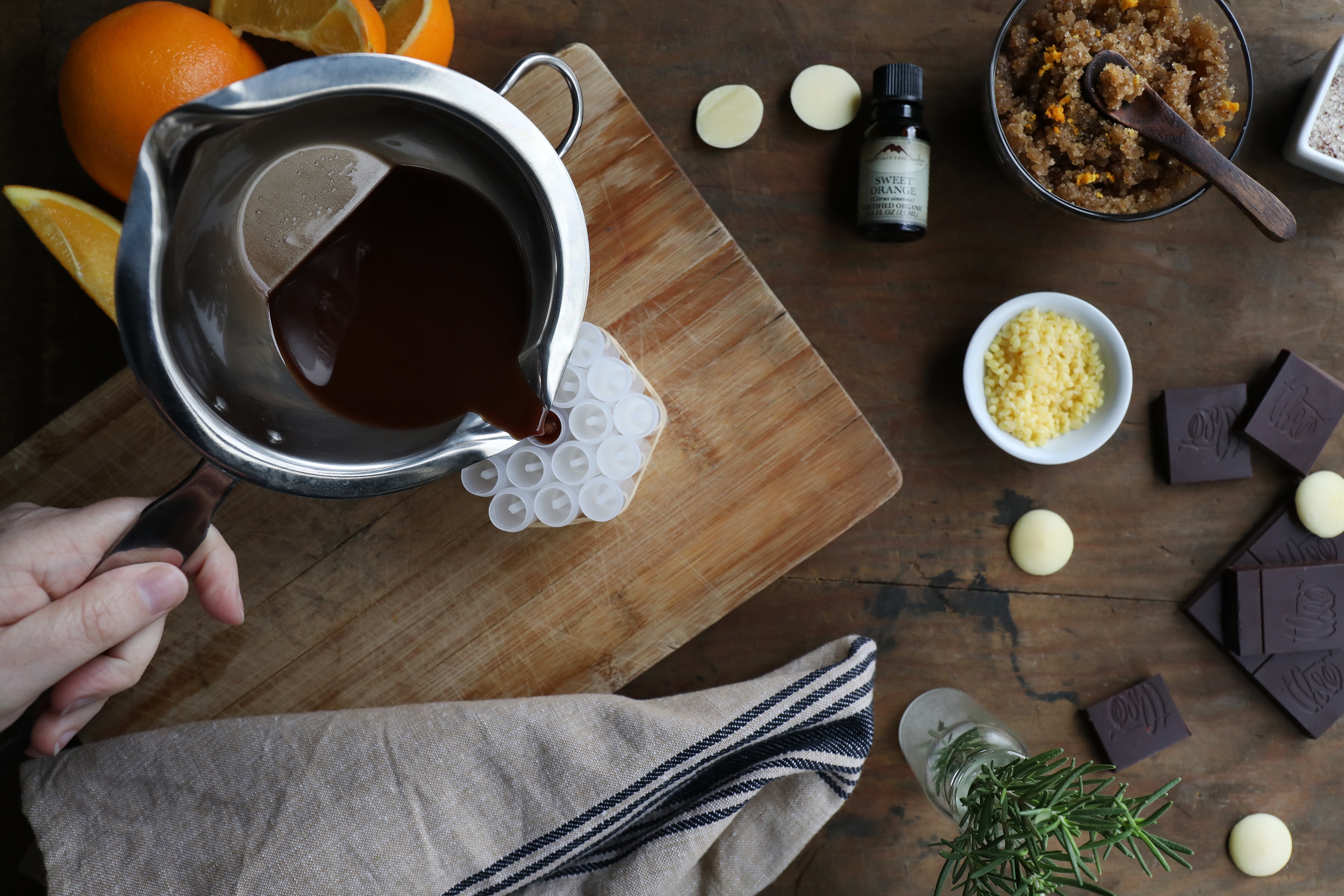 Hand pouring dark colored liquid into lib balm tubes with fresh citrus and rosemary and other ingredients surround it. 