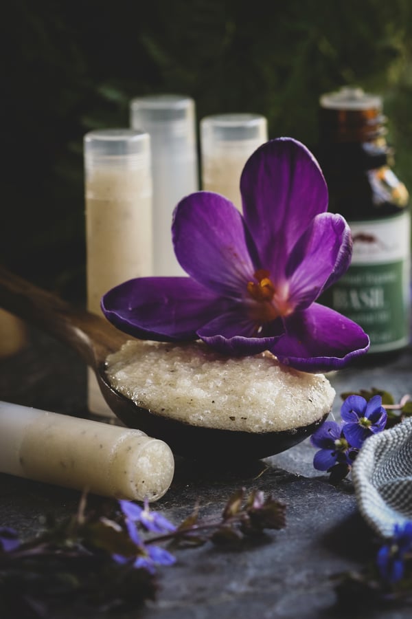 DIY recipe in a wooden spoon surrounded by filled lip balm tubes.  Scene is garnished with pretty purple flowers. 