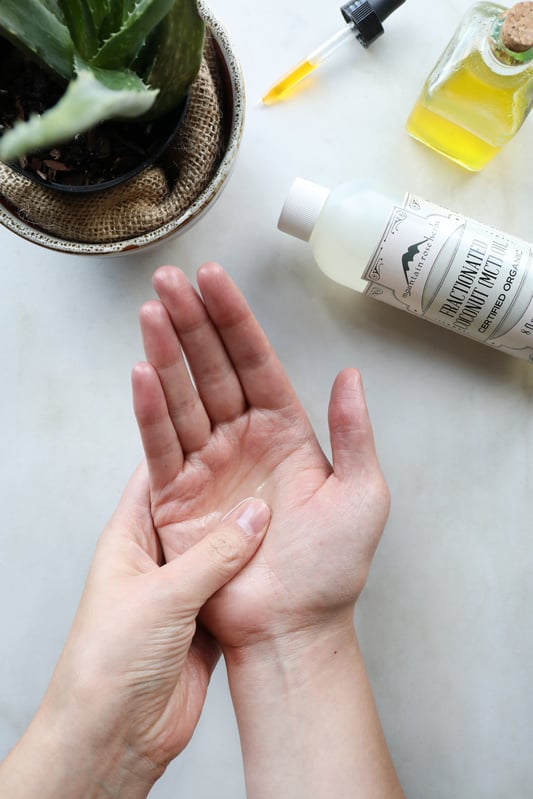 Hands applying oil to skin with a full bottle of fractionated coconut oil and plant in background. 