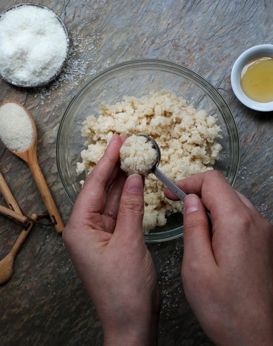 Hands scooping coconut macaroon mixture with other ingredients in background. 