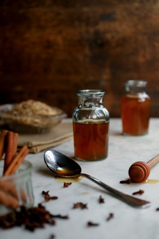 A spice bottle filled with a winter herbal syrup made from organic wellness herbs like echinacea purpurea, cinnamon, fennel, cloves, marshmallow, and ginger. A spoonful of herbal syrup made with licorice root and honey. 