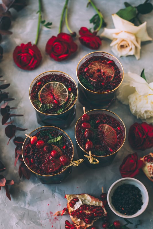 Holiday cocktails, or mocktails without alcohol, made with organic maqui berries and bilberries in trendy glass jars with gold rims and garnished with pomegranate seeds, fresh lime wheels, and cranberries.