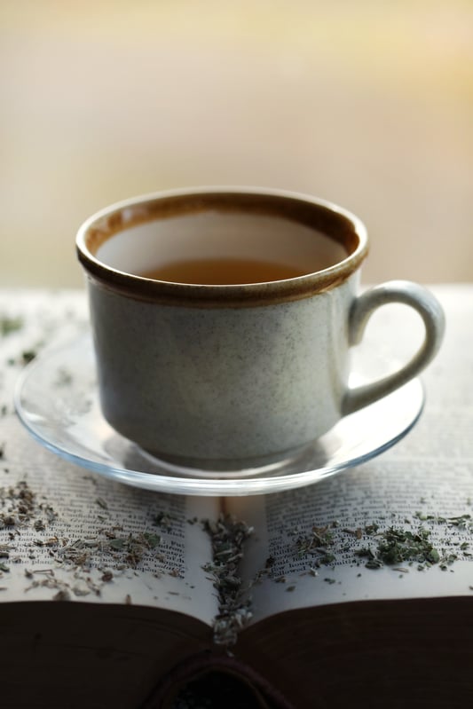 Vintage looking mug filled with golden liquid sitting on top of book with herbs spilled around