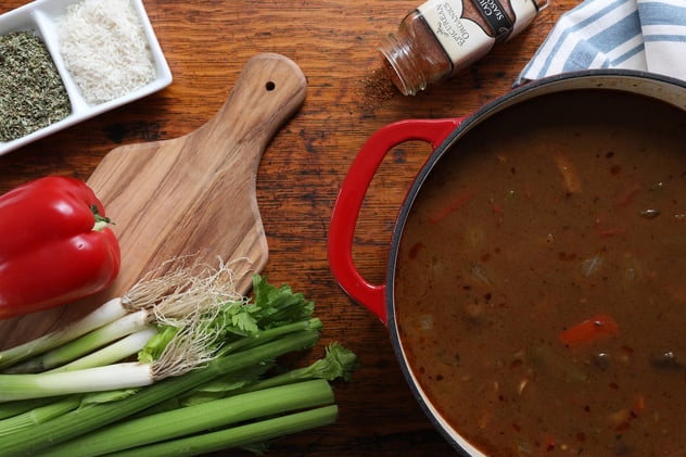 Vegetarian gumbo in pot with ingredients nearby on cutting board and spices and herbs nearby