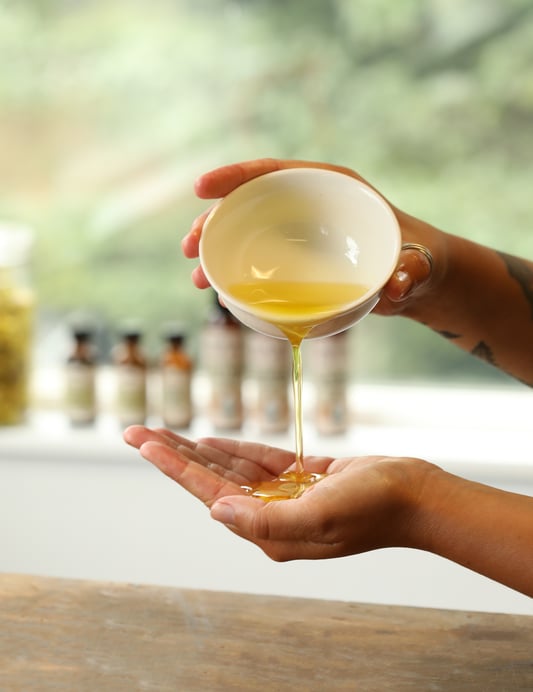 Golden massage oil is poured from a bowl to begin a relaxing massage. Several bottles of massage oils are arranged on a windowsill. 