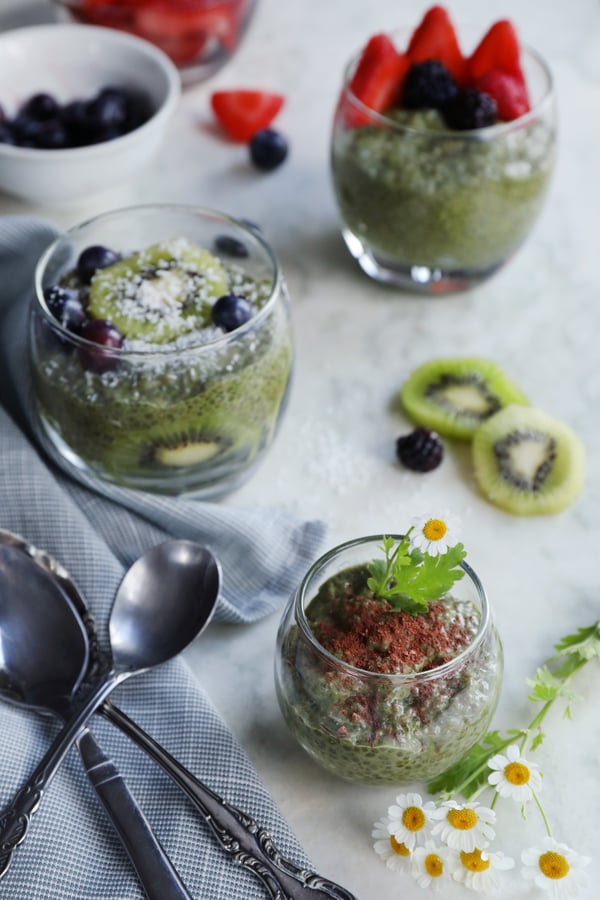 Clear cups of organic matcha chia pudding are garnished with fresh fruits and freshly picked feverfew flowers. Kiwis, blackberries, strawberries, and blueberries adorn the cups of green matcha chia pudding. 