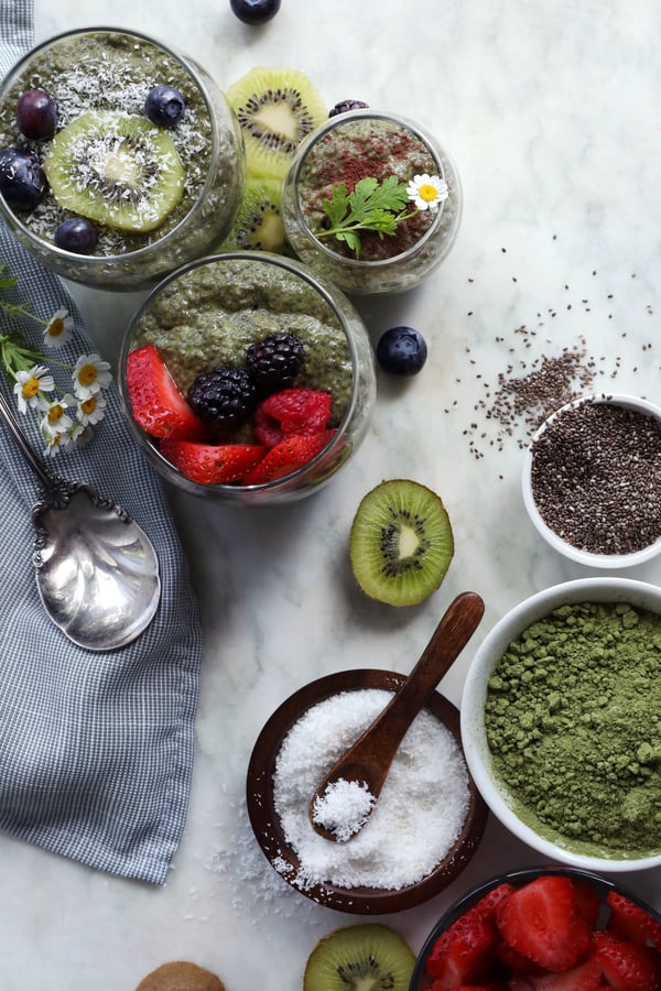 Ingredients are assembled to make matcha chia pudding. Organic coconut flakes, chia seeds, and matcha powder, along with fresh fruits and completed glasses of matcha chia pudding. 