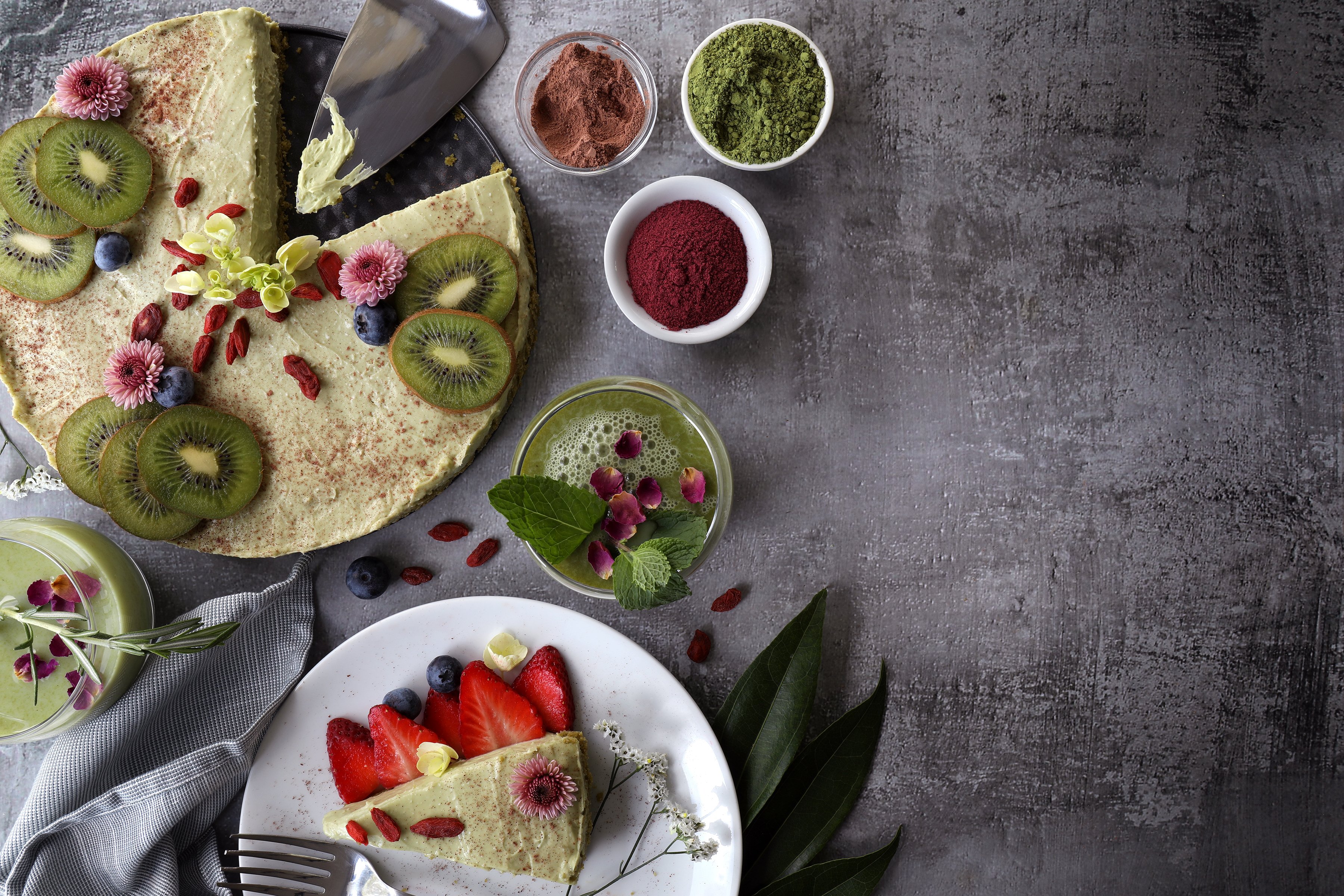 Beautiful matcha green tea cheese cake adorned with goji berries, fresh blueberries and kiwi fruit, and ornamental flowers with two glasses full of matcha latte. Arranged with strawberries, and bowls of colored powder on a gray slate background.