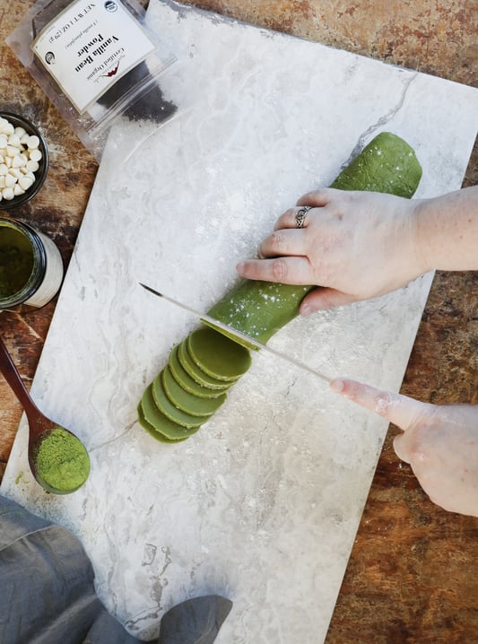 Hand slicing green cookie roll with vanilla powder and matcha tea displayed. 